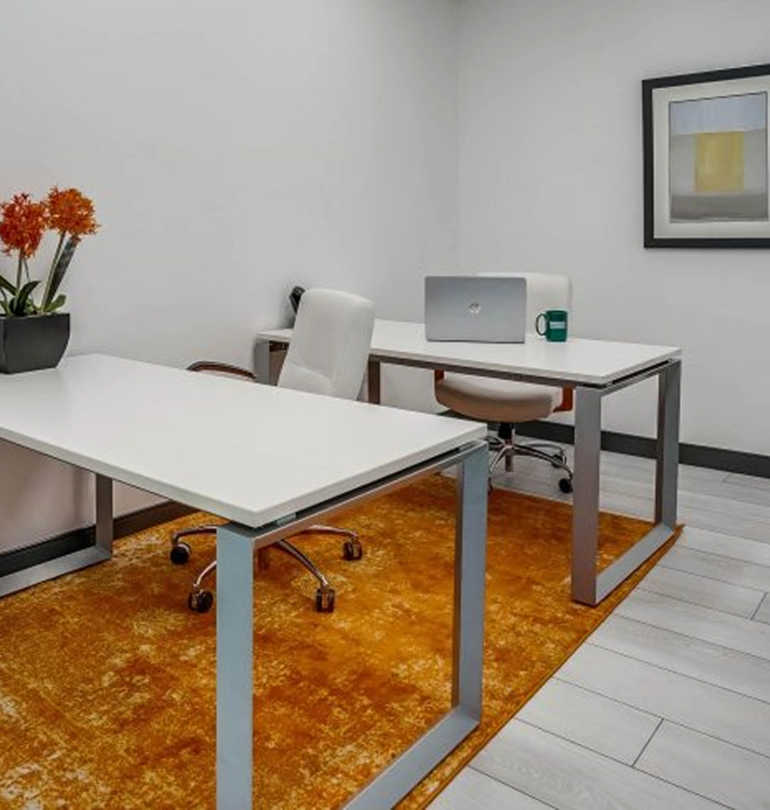 Modern office space with two white desks, white chairs, a laptop, green mug, orange flowers in a pot, and a framed abstract artwork on the wall. The floor features a bright orange rug and white tiles.
