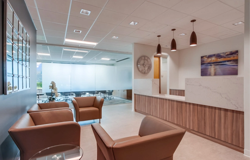 Modern office lobby with two brown armchairs, a wooden reception desk, pendant lights, a large wall clock, and framed artwork. Frosted glass walls enclose a meeting room in the background.