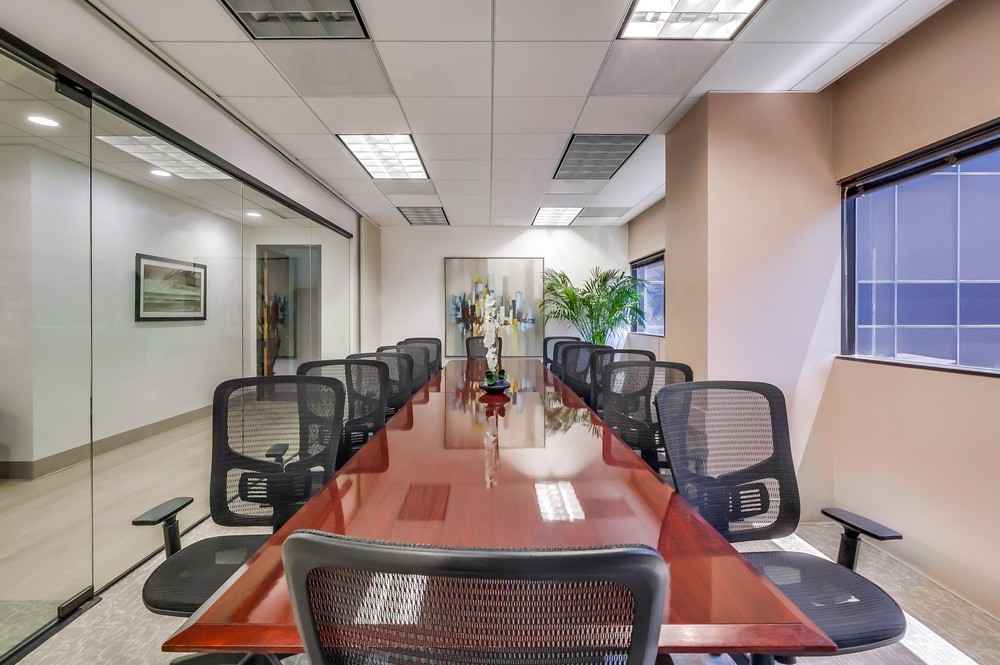 A modern conference room with a long wooden table, ten black mesh chairs, glass walls, artwork, a plant, and large windows letting in natural light.