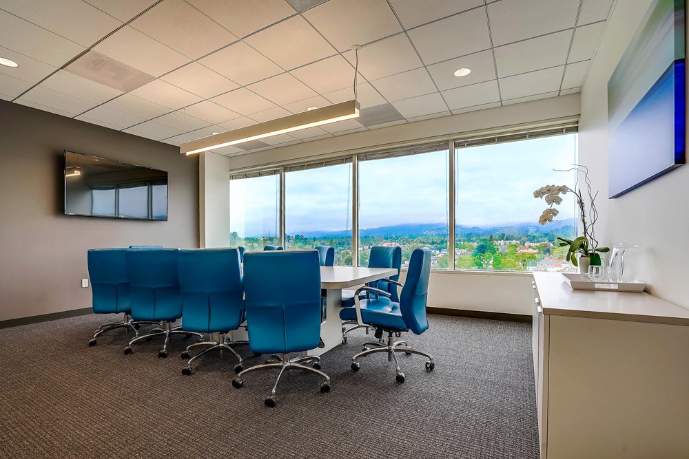 A modern conference room with blue chairs around a table, large windows showing a scenic view, a wall-mounted TV, and a cabinet with a plant and glassware.