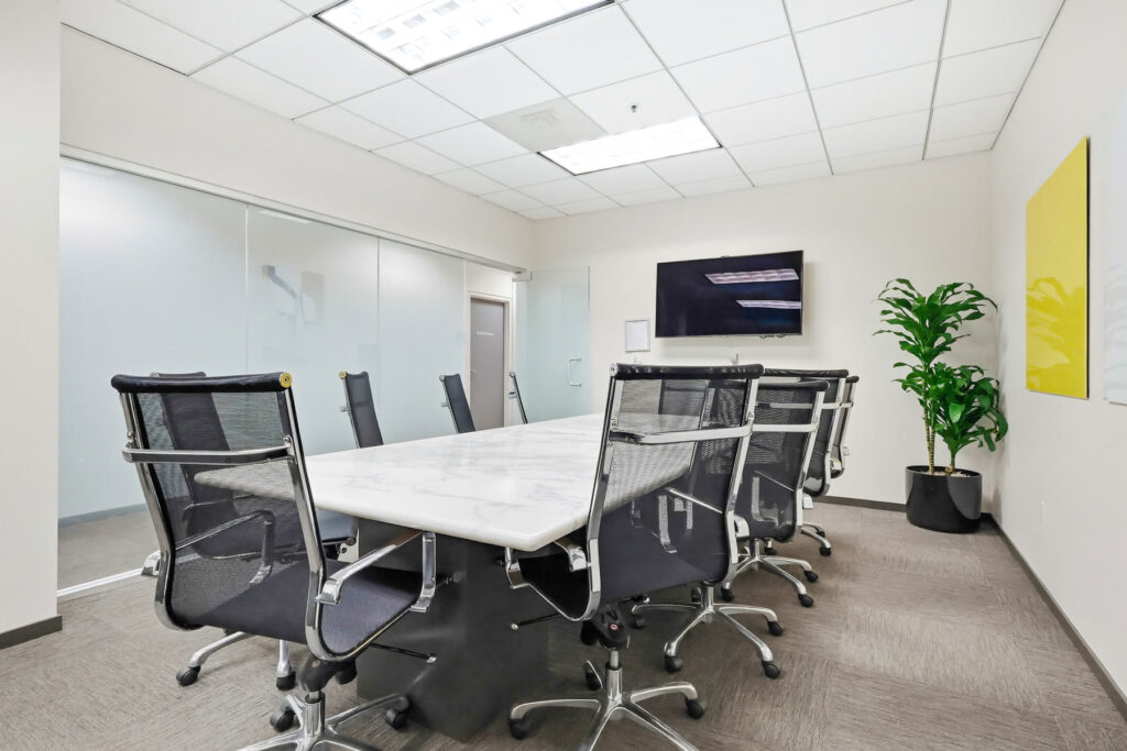 Modern conference room with a marble table, eight black office chairs, a wall-mounted screen, potted plant, glass wall, and bright overhead lighting. Artwork is hanging on the right wall.