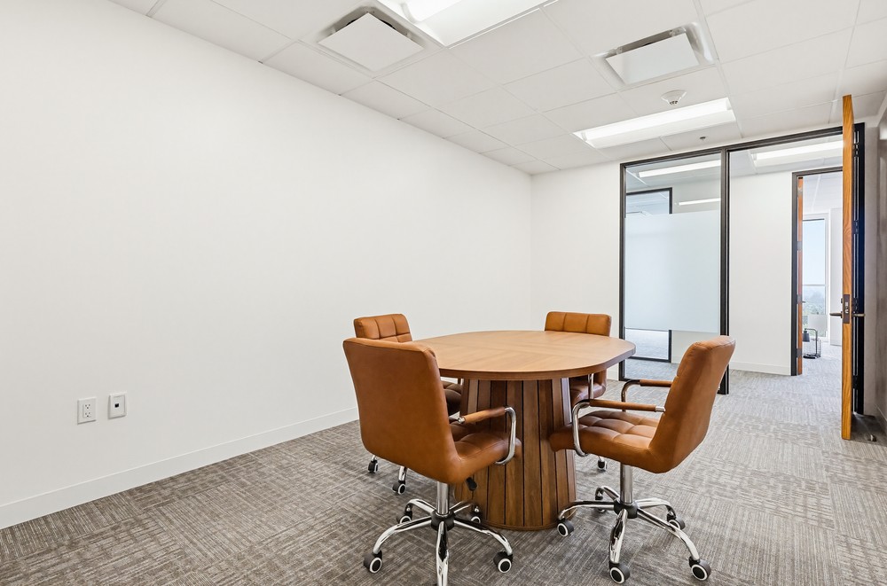 A small meeting room with a round wooden table, four brown leather chairs on wheels, beige carpet, white walls, and a glass door leading to another office space.
