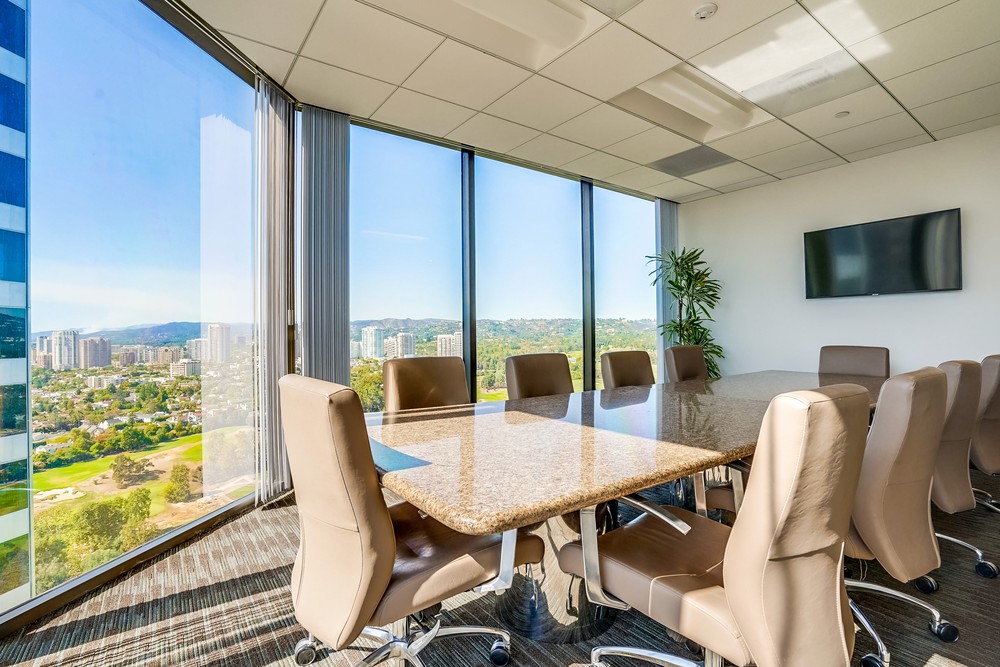 Modern conference room with a large window view of a cityscape, beige chairs around a marble table, wall-mounted TV, plant in corner, and bright natural light.