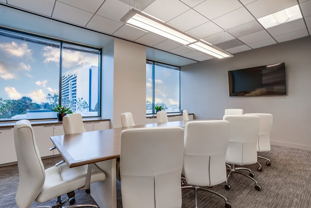 Modern conference room with a large wooden table, eight white chairs on wheels, a wall-mounted TV, two large windows with city views, and potted plants on the windowsills.