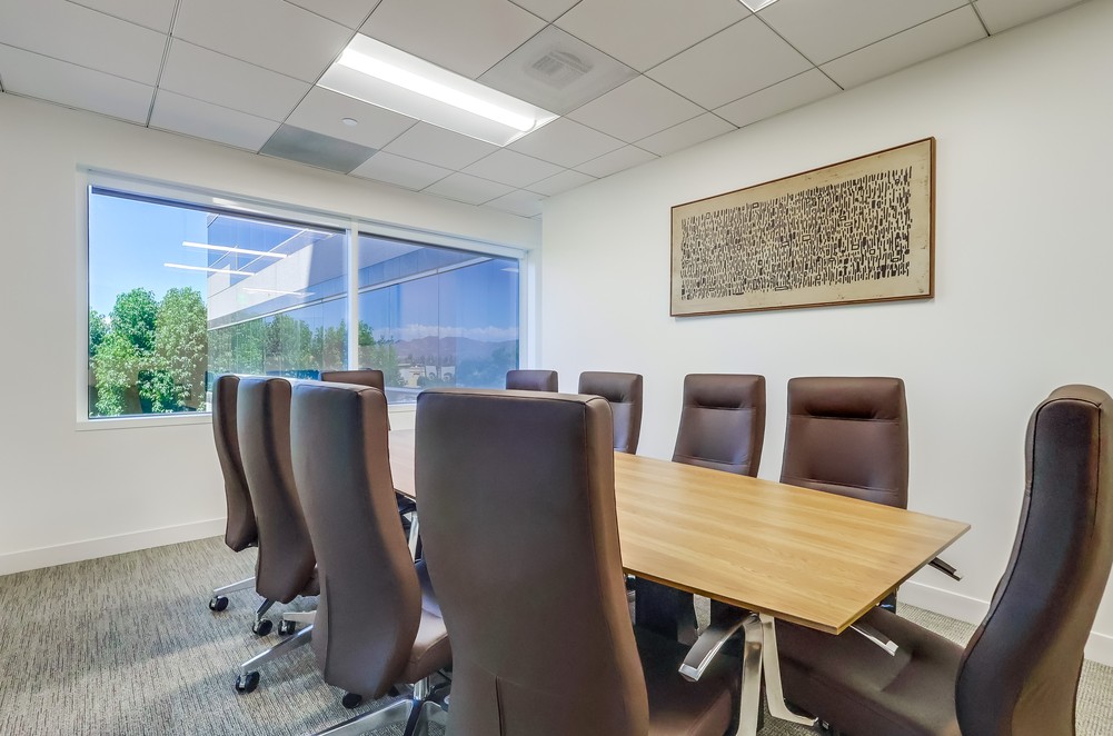 A modern conference room with a wooden table, eight brown chairs, a large window with outdoor views, and abstract artwork on the wall.