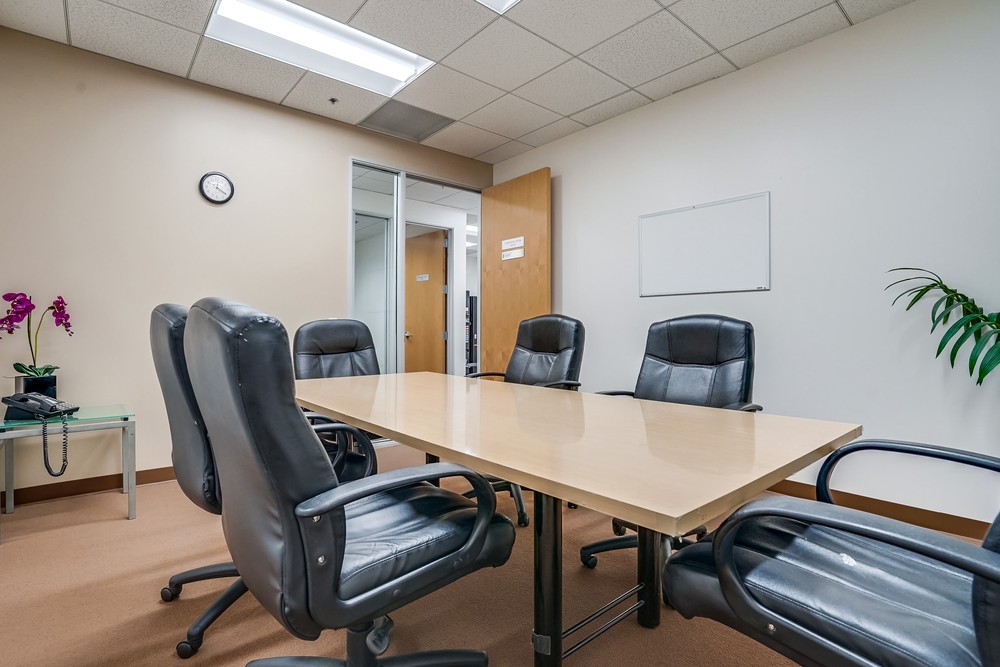 A small conference room with a table, five black chairs, a wall clock, a whiteboard, a plant, and a side table with a phone and purple flowers. The doors are open, showing part of a hallway outside.