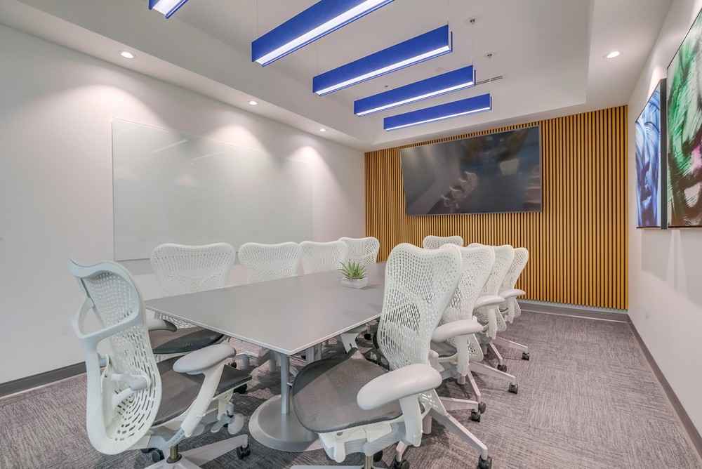 Modern conference room with a long table, ten white ergonomic chairs, a large wall-mounted screen, a whiteboard, and minimalist decor including a small plant on the table.