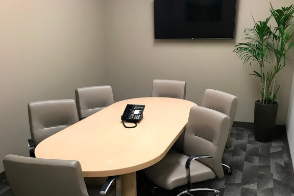 A small meeting room with six beige chairs around an oval table, a phone on the table, a wall-mounted TV, a potted plant in the corner, and gray carpeted floor.