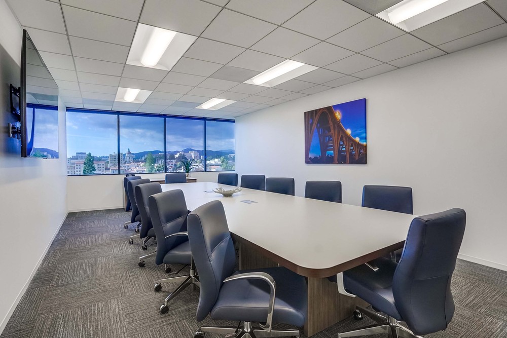 Modern conference room with a long table, twelve blue chairs, large window with city view, wall-mounted TV, and a framed photo of a bridge on the white wall.