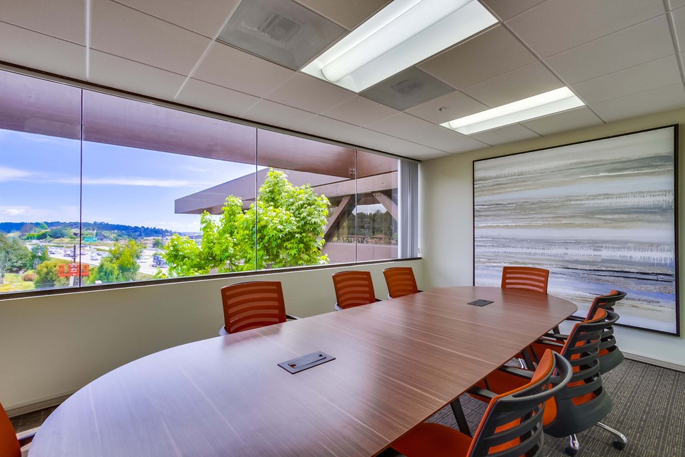 Modern conference room with a long wooden table, orange chairs, large window with a scenic outdoor view, and a large abstract painting on the wall.