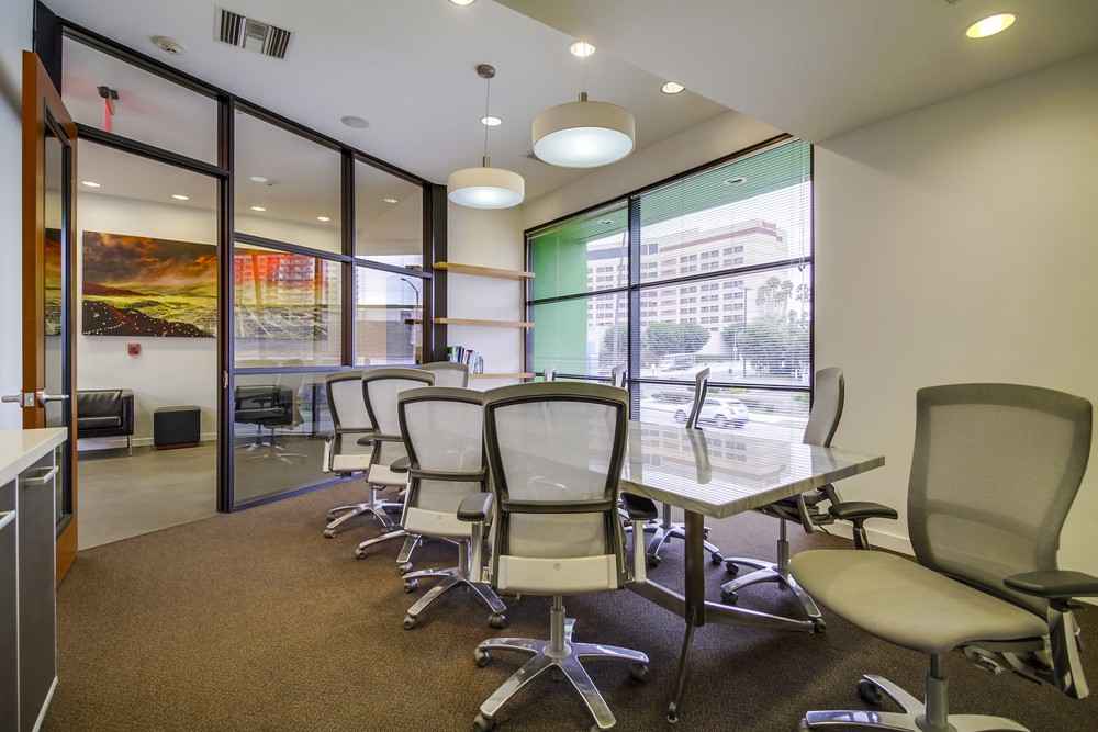Modern conference room with a long table, several gray office chairs, large windows, hanging lights, and glass walls. City buildings are visible outside, and a small sitting area is next to the meeting room.