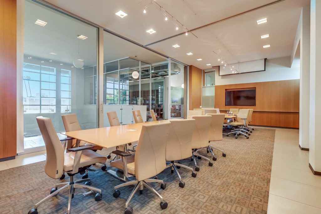 Modern conference room with a long wooden table, beige swivel chairs, glass walls, and natural light coming through large windows.