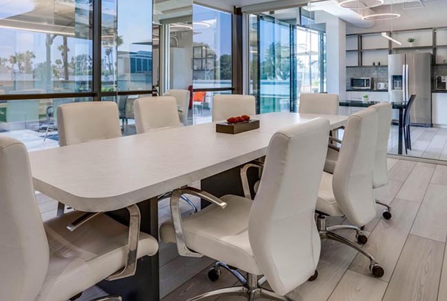 A modern conference room with a white table, eight white chairs on wheels, glass walls, and a view into a kitchen area. A small tray with red apples sits in the center of the table.