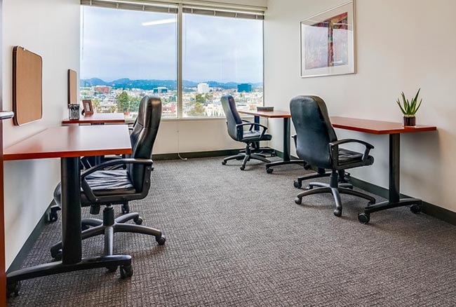 Modern office with three desks and office chairs, a large window with a city view, a framed picture on the wall, and a small plant on one desk. The room has neutral colors and natural light.