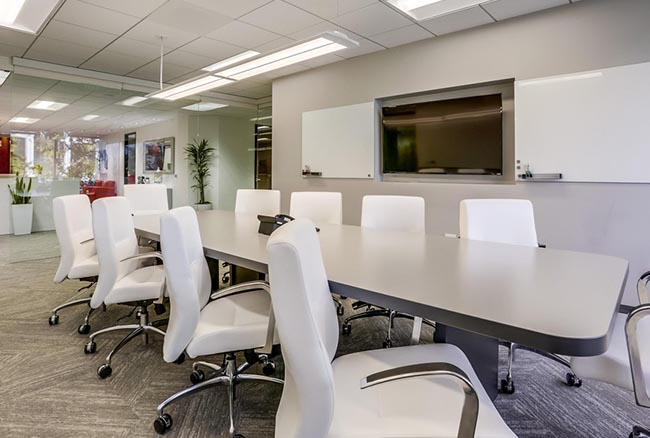 Modern conference room with a long table, white chairs, two whiteboards, a large monitor, bright lighting, and glass walls. Green plants and office decor visible in the background.