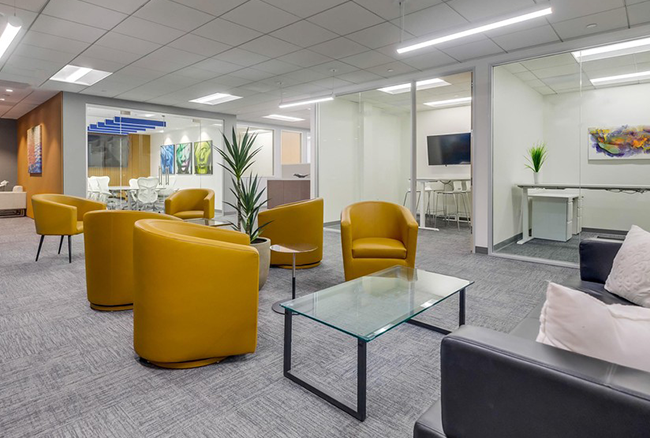 Modern office lounge with yellow chairs, glass coffee table, gray sofas, potted plants, and glass-walled meeting rooms in the background, with abstract art on the walls and gray carpeted floors.