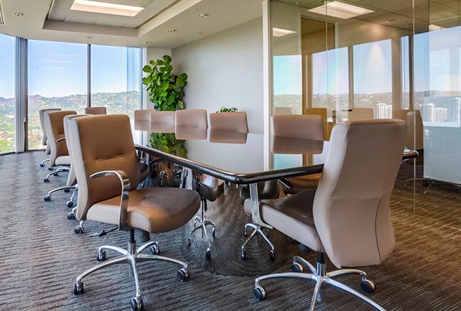 Modern conference room with a long glass table, beige office chairs on casters, a large window with a city view, glass walls, and a potted plant in the corner.