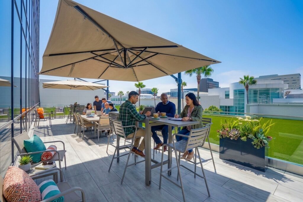 People sit at tables on a modern rooftop patio, socializing and eating under large umbrellas. Green plants and city buildings are visible in the background under a clear blue sky.