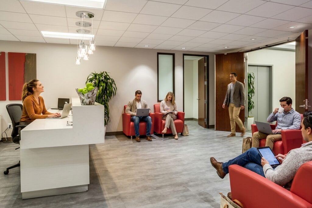Modern office lobby with a receptionist at a desk. Four people sit on red chairs using laptops and phones, while one person enters through an open door. There are plants and abstract art on the wall.