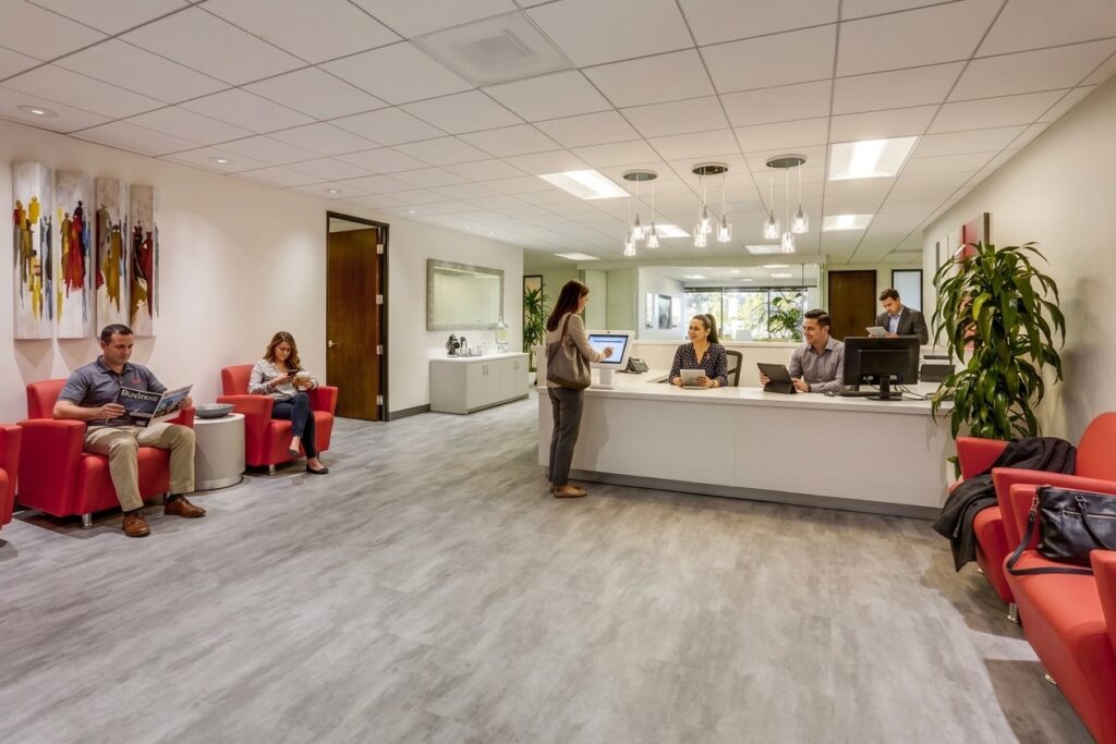 Modern waiting room with red chairs, people sitting and reading, a receptionist desk with staff assisting a woman, and bright lighting. Plants and artwork decorate the space.