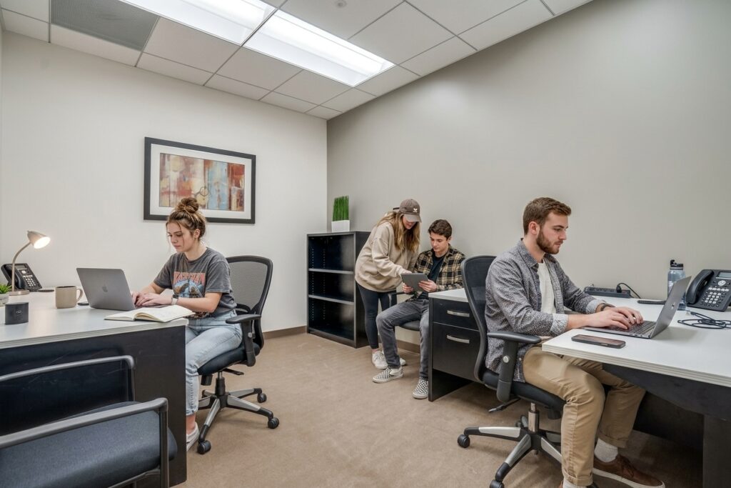 Four people work in a modern office. Two sit at desks using laptops, while two others stand nearby discussing something on a tablet. The room has neutral walls and a framed picture.