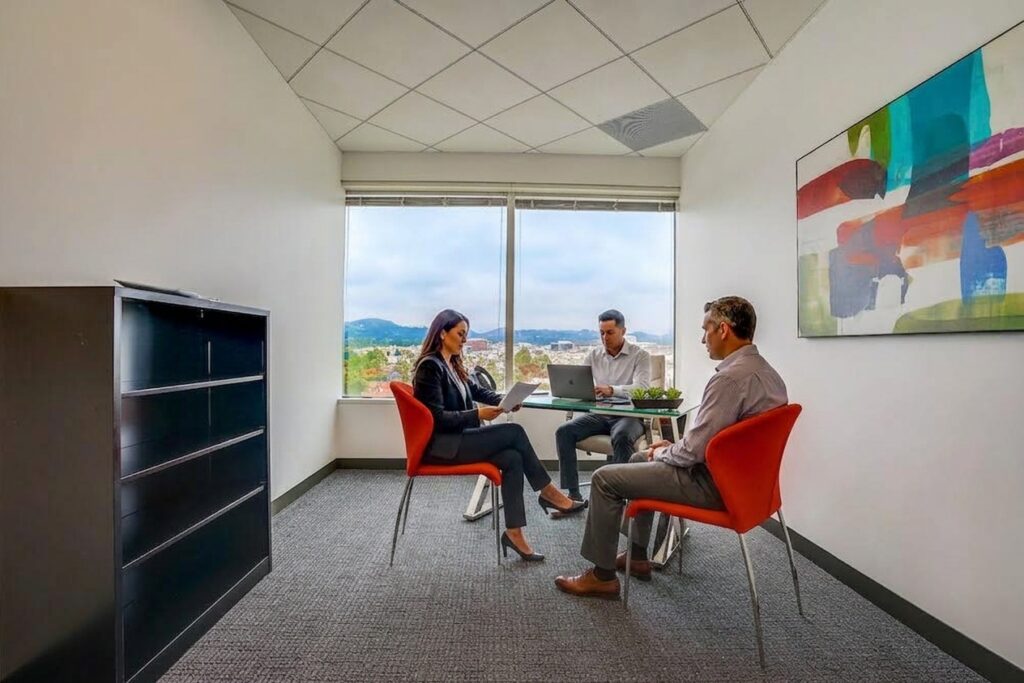 Three people sit on orange chairs around a small table in a modern office. Large window with scenic view, colorful abstract painting on the wall, and a black filing cabinet are visible.