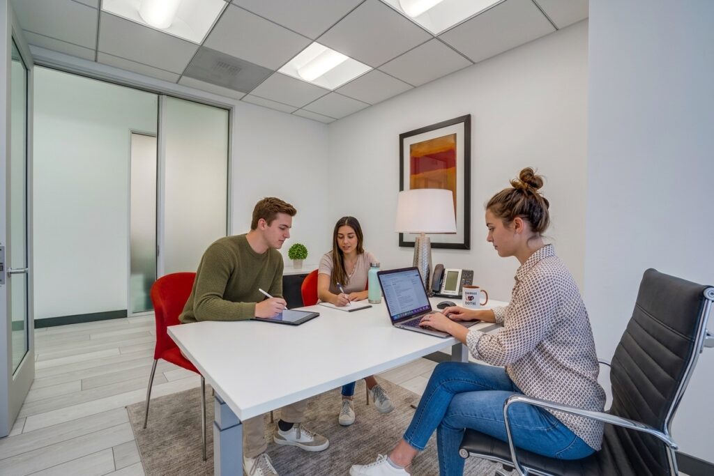 Three people are working together in a modern office, seated around a white table with laptops, notebooks, and pens. The office has white walls, a lamp, artwork, and red chairs.