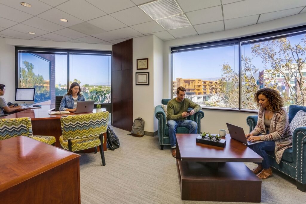 Four people work in a modern office with large windows. Two sit at a wooden desk with laptops, while two others sit on chairs around a coffee table, also using laptops. Trees and buildings are visible outside.
