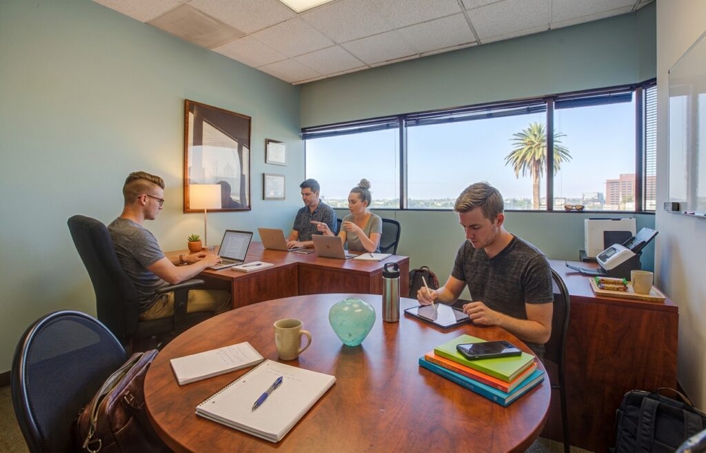 Four people work at desks in a bright office with large windows, laptops, notebooks, coffee mugs, and a palm tree visible outside.