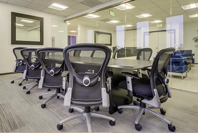 Modern conference room with a long black table, mesh office chairs, glass walls, and a large mirror on the wall. Bright, neutral colors and natural light create an open and professional atmosphere.