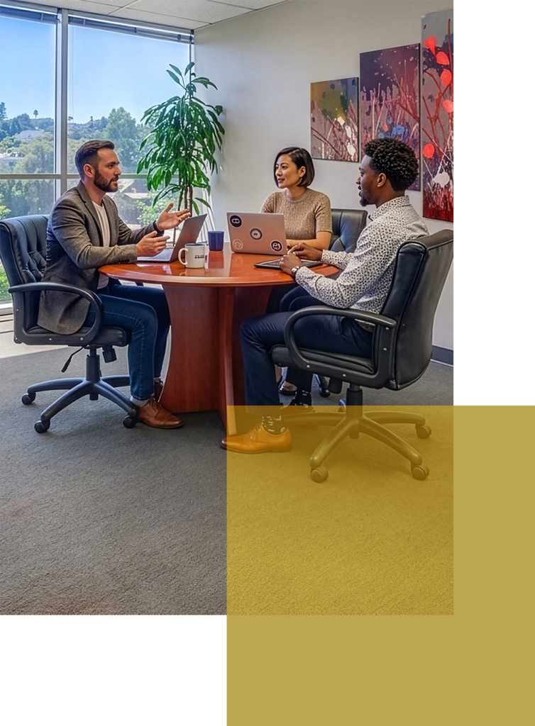 Three people sit around a round table in an office, discussing work. Two have laptops open. Large windows, plants, and colorful wall art are visible in the background.
