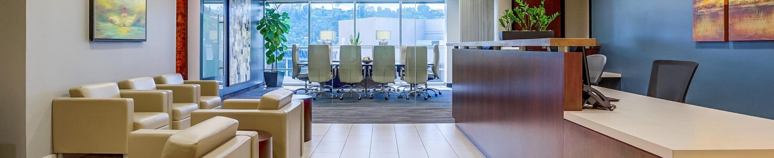 Modern office lobby with beige armchairs, a reception desk, potted plants, wall art, and a glass-walled conference room with white chairs and a view of trees and buildings outside.