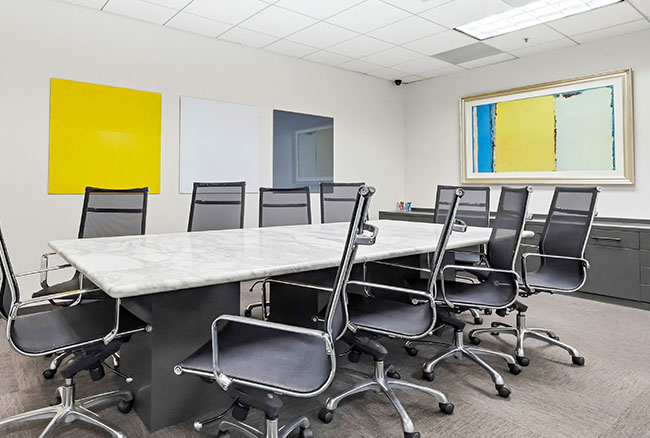 A modern conference room with a marble table, ten black office chairs, and abstract art on white walls. The room is well-lit with ceiling lights and has a neutral carpeted floor.