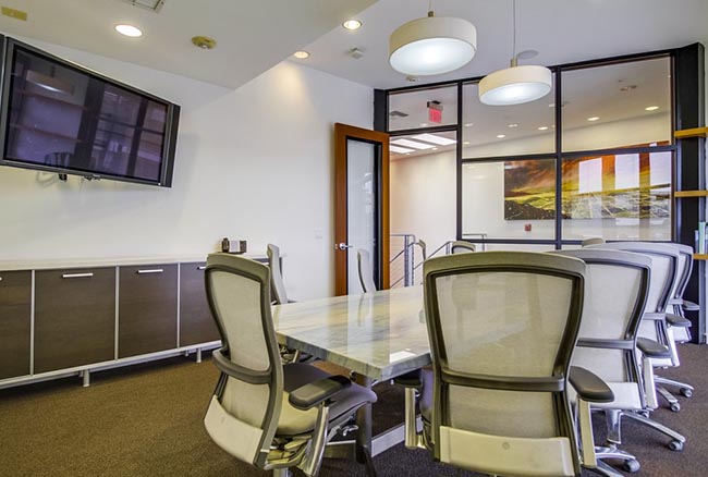 Modern conference room with a rectangular table, several mesh office chairs, a wall-mounted TV, cabinets, overhead lights, and large glass windows revealing art and natural light in the adjacent space.
