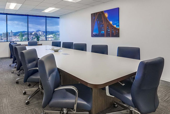 Modern conference room with a long white table, twelve blue chairs, large windows showing a city view, and a framed photo of a lit bridge on the white wall.