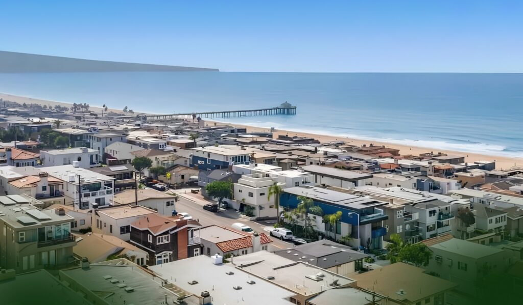 A coastal neighborhood with many houses, a sandy beach, and a long pier stretching into the ocean under a clear blue sky.