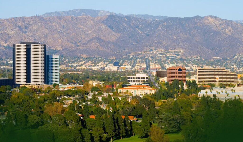 A cityscape featuring tall office buildings, residential areas, and lush green trees, with mountains in the background under a clear sky.