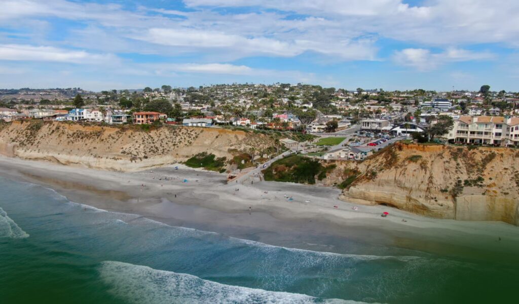 Aerial view of a sandy beach with gentle waves, backed by tan cliffs and stair access. Houses and greenery are visible on the cliffs under a partly cloudy sky.
