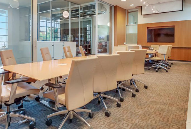 A modern conference room with beige chairs around two wooden tables, a large monitor on the wall, glass partitions, and carpeted floor. The room is brightly lit with natural and overhead lighting.