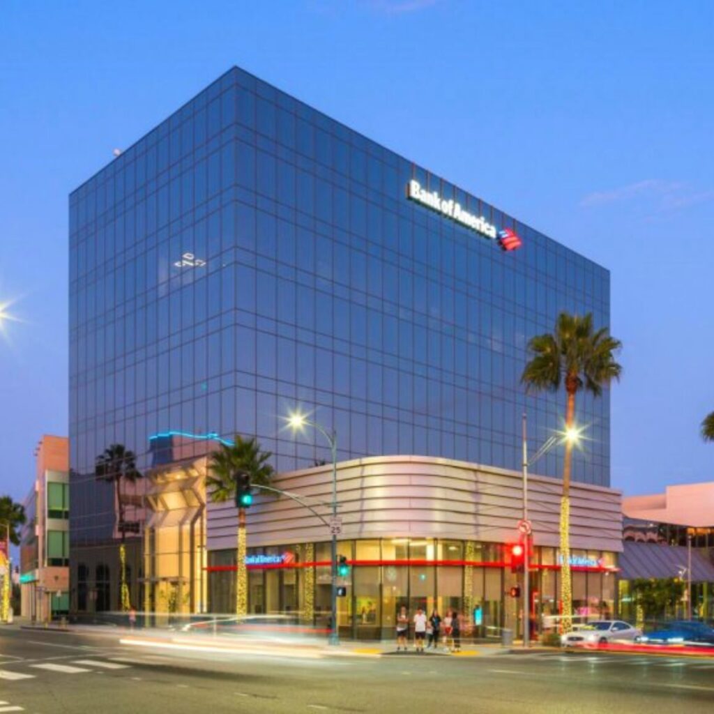 A glass office building with a Bank of America sign at the top, palm trees in front, and city lights and traffic at dusk.