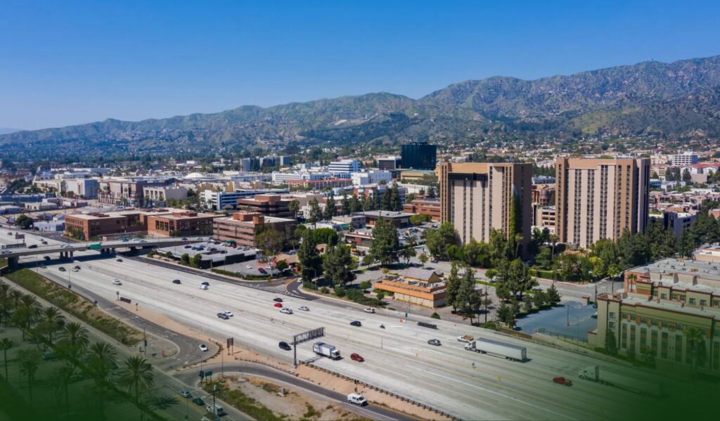 A wide freeway runs through a city with tall buildings, trees, and mountains in the background under a clear blue sky.
