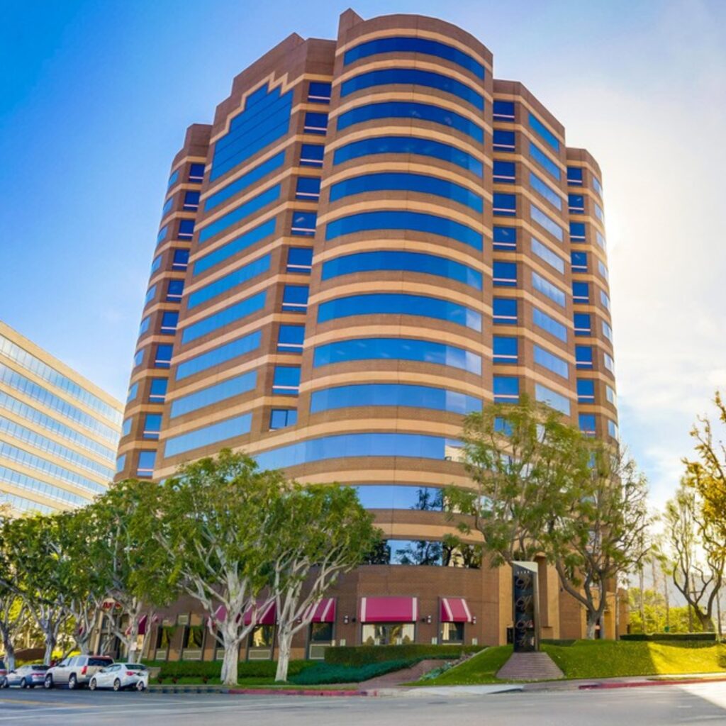 A tall, modern office building with brown and blue glass exterior, surrounded by trees and parked cars, under a clear sky.