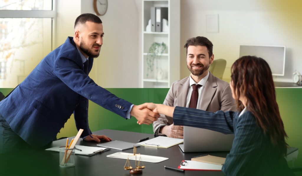 Three people in business attire sit at a desk in an office. Two of them, a man and a woman, are shaking hands while the third man smiles and watches. Laptops, papers, and office items are on the desk.