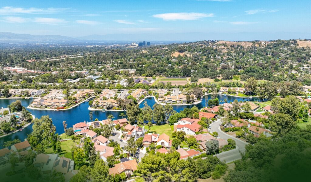 Aerial view of a suburban neighborhood with red-roofed houses, winding streets, lakes, and abundant green trees under a blue sky.