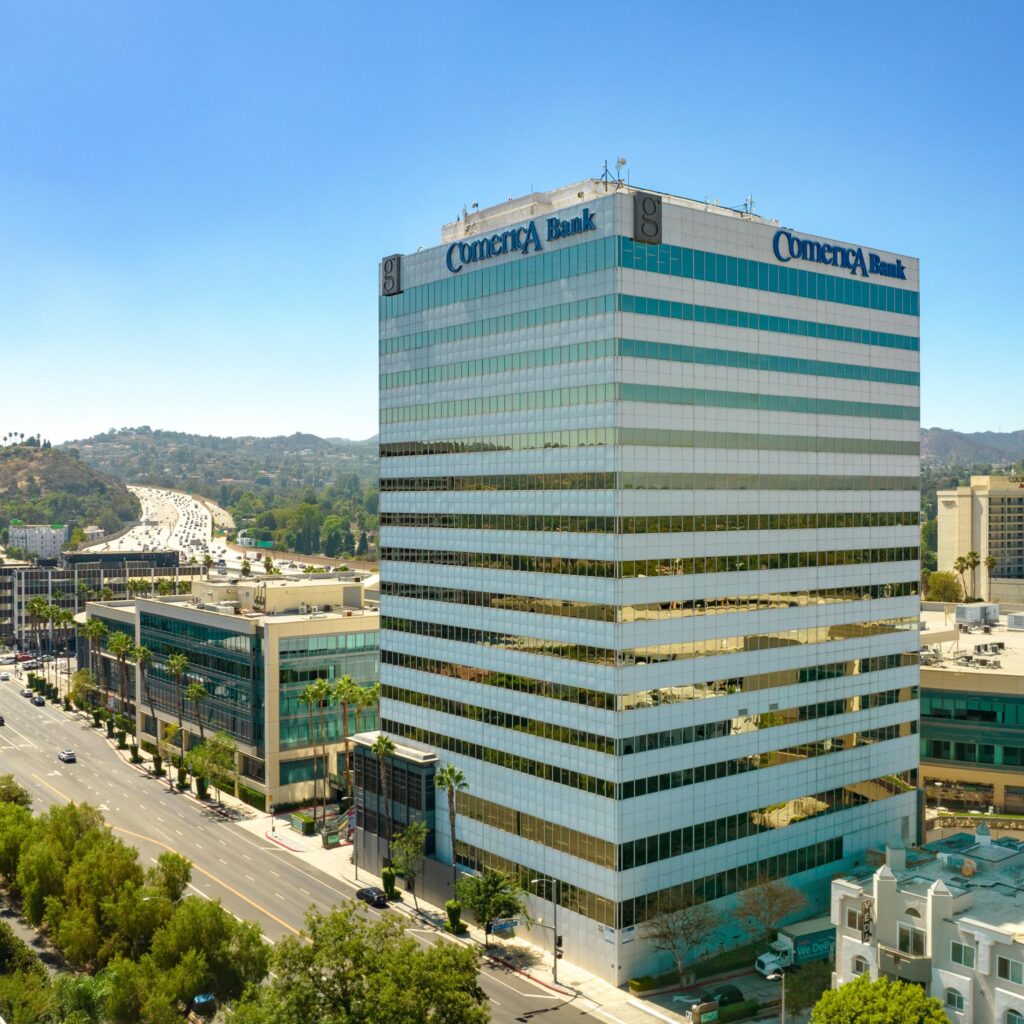 A tall office building with Comerica Bank signage, surrounded by other buildings and trees, stands next to a busy road under a clear blue sky.