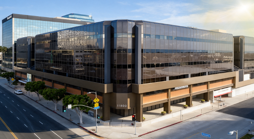 An aerial view of a modern office complex at 11500 Olympic Boulevard, featuring dark glass facades, brown structural trim, and a street-level entrance with a City National Bank sign. The street corner includes traffic lights and green trees.