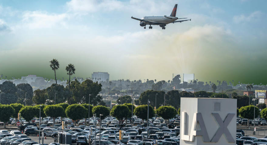 An airplane descends over a crowded parking lot near the LAX sign, with palm trees and buildings in the background under a partly cloudy sky.