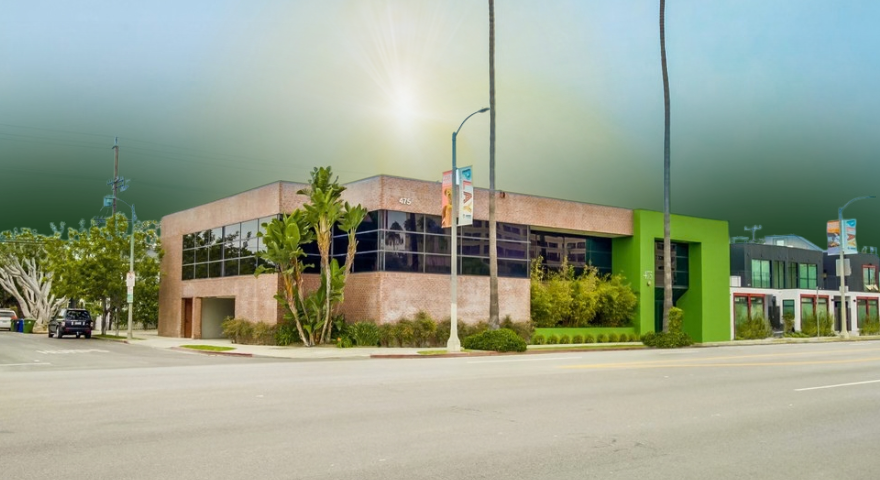 A modern two-story brick building with green accents, large windows, and surrounding plants sits on a quiet street under a bright sky with sunlight shining above.