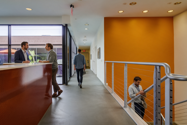 Two people talk at a modern office reception desk. Another person walks down the hallway, and one person is on the stairs looking at a phone. The space features large windows and an orange accent wall.