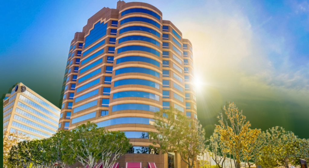 A tall, modern office building with curved rows of windows stands against a bright sky, with sunlight shining through scattered clouds and trees in the foreground.